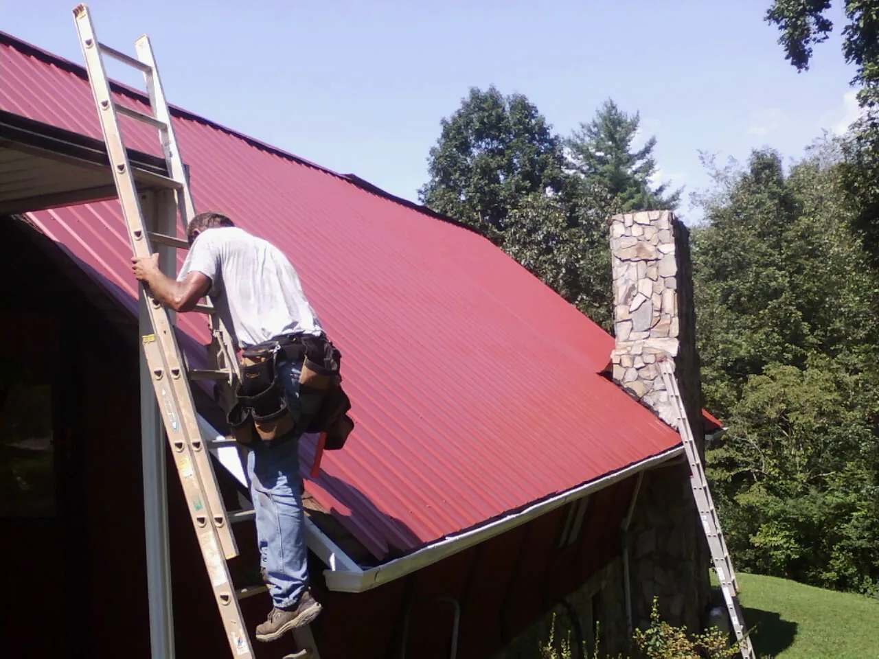Roofer on a ladder beside a red metal roof and stone chimney — Soffit & Fascia Repair by Roofing Brothers in Harvey